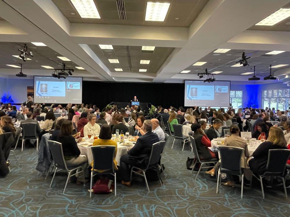 People seated at round tables in a ballroom, listening to a presentation from a speaker on a stage at the 2023 Behavioral Health Convening.